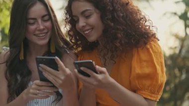 Happy calm young girls friends looking at their smartphones and smiling while sitting in the park