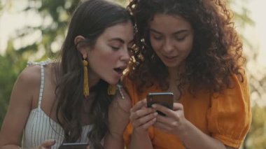 Attractive young girl looking at her friend's smartphone and becoming surprised while sitting in the park