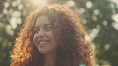 Attractive flirty young redhead curly woman smiling and looking at someone to the side while standing in the park