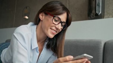 Evdeki kanepede yatarken gülümseyen ve akıllı telefon kullanan neşeli genç iş kadınının yakın görüntüsü