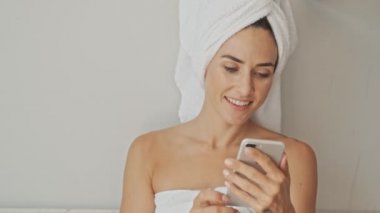 Banyo yaptıktan sonra beyaz havlu giyen ve sabah evde dinlenirken akıllı telefon kullanan alımlı genç bir kadın.