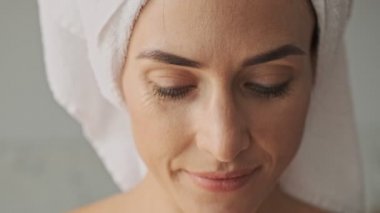 Kafasında beyaz havluyla, banyodan sonra evde dinlenirken gülümseyen ve kameraya bakan çekici, mutlu genç bir kadın manzarası.