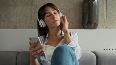 Çekici, genç bir kadın, gözleri kapalı gülümsüyor ve bir kanepede otururken kablosuz kulaklıkla müzik dinliyor.