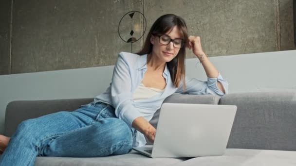 魅力的な若いビジネス女性の笑顔とノートパソコンで作業しながら 自宅でソファに座って — ストック動画