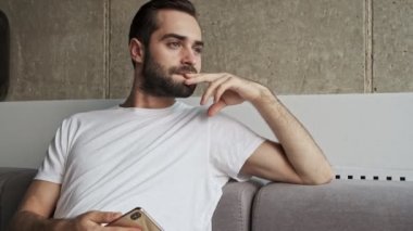 Akıllı telefonlu yakışıklı esmer adam üzgün hissediyor ve evdeki kanepede otururken pencereye bakıyor.