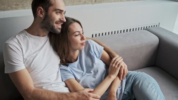 魅力的な笑顔若い素敵なカップル抱擁の側面ビューと自宅でソファに座っている間 ウィンドウを見て — ストック動画