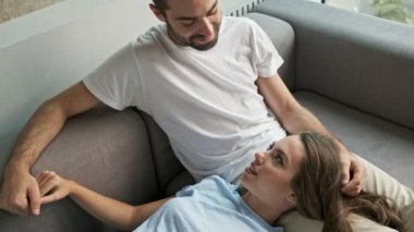 Attractive young woman lying on her boyfriend's legs and talking with him while relaxing on couch at home