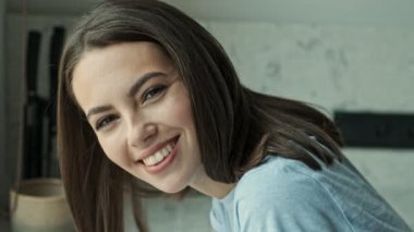 Mutfakta otururken gülümseyen ve kameraya bakan güzel, esmer bir kadının yan görüntüsü.