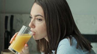 Mutfakta oturup meyve suyu içen ve gülümseyen hoş, genç esmer kadının yan görüntüsü.