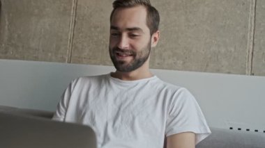 Evdeki kanepede otururken laptopta gülümseyen ve sohbet eden yakışıklı, mutlu esmer adamın yakın görüntüsü.