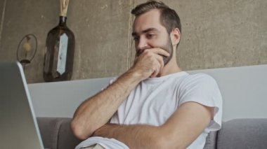Evdeki kanepede otururken dizüstü bilgisayarda çalışan konsantre genç esmer adam.
