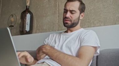 Upset serious young brunet man working on laptop while sitting on sofa at home