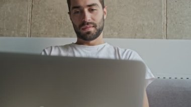 Yakışıklı, ciddi, esmer bir adamın dizüstü bilgisayarda çalışmasını ve evdeki kanepede otururken mutlu olmasını yakından izleyin.