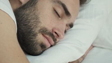 Yakışıklı, gülümseyen esmer adamın yakın görüntüsü gözlerini açıyor ve sabah yatakta yatarken uyanıyor.