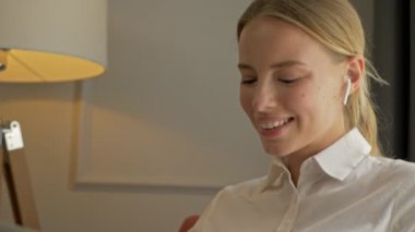 Cheerful cute young businesswoman chatting on smartphone and drinking wine while working on laptop sitting in armchair near the window