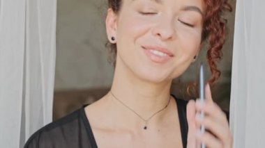 İpek bornozlu, mutlu, genç ve güzel bir kadının sabahları pencereye bakarken akıllı telefondan konuşmasının yakın görüntüsü.