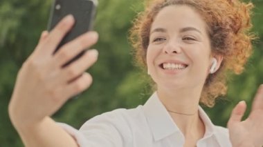 Beyaz tişörtlü, güzel, mutlu, genç bir kadın. Parktaki akıllı telefondan bir video görüşmesi yaparken gülümsüyor ve el sallıyor.