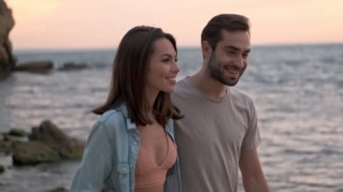 Çekici, mutlu, genç bir çift, deniz kenarında yürürken gülümsüyor ve sarılıyor.