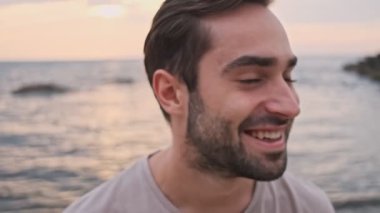 Yakışıklı, neşeli, sakallı adamın yakın görüntüsü. Gülümseyen ve saçlarını deniz kenarında dinlenirken elleriyle düzelten adam.