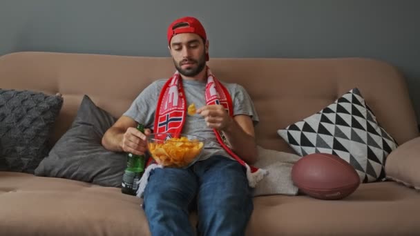 帽子をかぶった男性のファンがビールを飲みながらソファに座ってアパートでスポーツを見ている間にチップを食べるハンサムな髭 — ストック動画