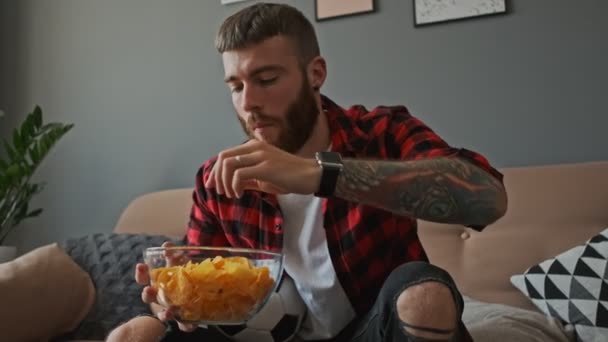 坐在沙发上和公寓里看体育活动时 全神贯注的大胡子男粉丝正在吃薯片 — 图库视频影像