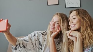 İki neşeli genç kız arkadaş akıllı telefondan selfie çekiyorlar ve evdeki kanepede otururken barış jesti yapıyorlar.
