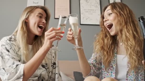 两个年轻快乐的女孩朋友一边喝着香槟 一边坐在家里的沙发上享受着这一刻 — 图库视频影像