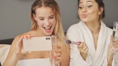 İki güzel kız arkadaş evde şampanya içerken akıllı telefondan selfie çekiyorlar.