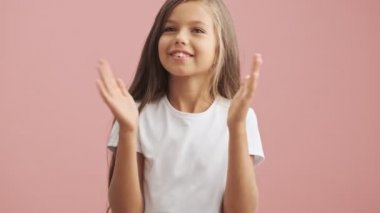 Gülümseyen küçük kız el çırpıyor ve pembe arka planda tek başına kameraya bakıyor.