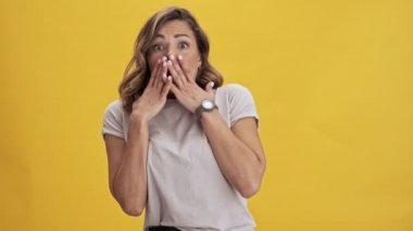 Kırmızı dudaklı, korkmuş genç bir kadın, sarı arkaplanda kameraya bakarken elleriyle yüzünü kapatıyor.