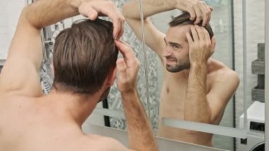 Banyodaki aynaya bakarken saçına dokunan, sakalsız, yakışıklı, genç bir adamın arka görüntüsü.