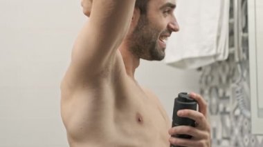 Yakışıklı, sakallı, sakalsız, gülümseyen ve banyodaki aynaya bakarken koltukaltına deodorant sıkan adamın yan görüntüsü.