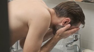 Gülümseyen, sakallı, genç ve sakalsız bir adamın banyo lavabosunda yüzünü yıkamasının yan görüntüsü.
