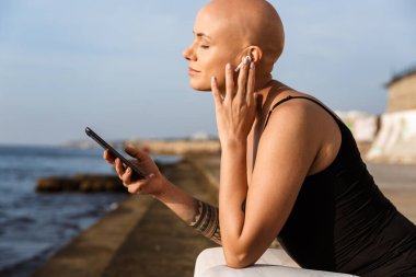 Otururken cep telefonu ve kulaklık kullanan gevşemiş kel kadın görüntüsü