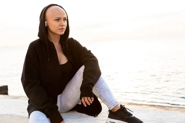 Imagen de una joven mujer calva usando auriculares mientras está sentada en el paseo marítimo —  Fotos de Stock