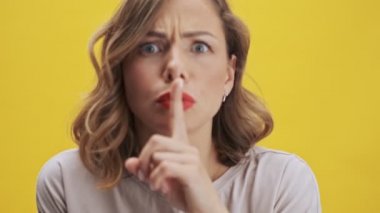 Kırmızı dudaklı güzel bir kadın, öfkeli duygularıyla sessiz bir jest yapıyor ve sarı arka plandan kameraya bakıyor.