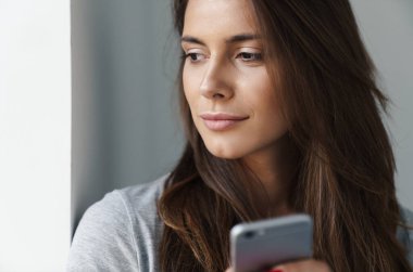 Gri arka planda gri bir duvara yaslanmış, cep telefonu kullanan genç ve güzel bir kızın yakın çekimi.