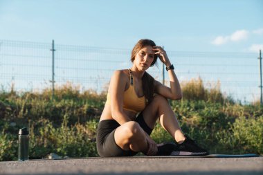Spor giyim giymiş esmer bir kadın oyun sahasında oturuyor.
