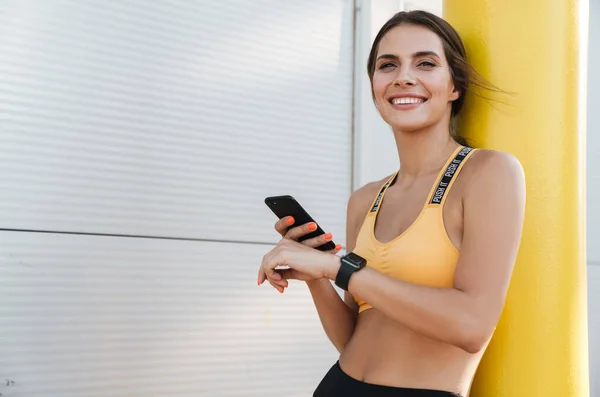 Obrázek radostné ženy ve sportovním oblečení držící mobilní telefon venku — Stock fotografie