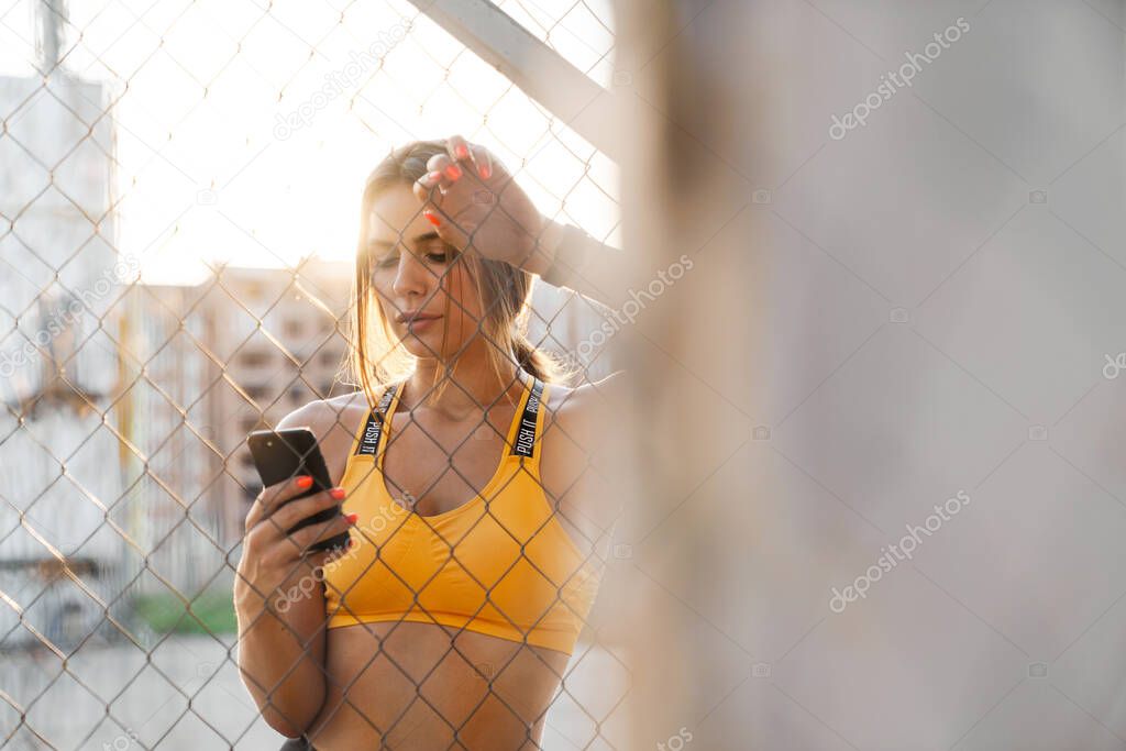 Image of beautiful woman holding cellphone by metal fence outdoo