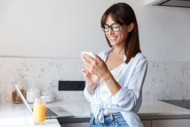 Güzel, neşeli, mutlu iş kadını evde, mutfakta cep telefonu kullanarak dizüstü bilgisayar kullanıyor..
