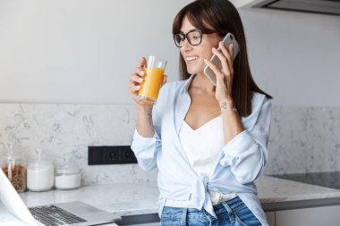 Mutfaktaki memnun iş kadını cep telefonuyla konuşan dizüstü bilgisayarla meyve suyu içiyor..