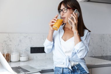Mutfaktaki memnun iş kadını cep telefonuyla konuşan dizüstü bilgisayarla meyve suyu içiyor..