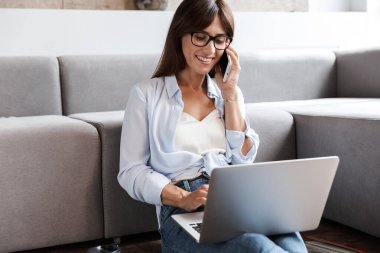 Oturma odasındaki koltukta oturan iş kadını cep telefonuyla konuşan dizüstü bilgisayarla konuşuyor..