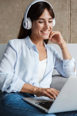 Evdeki kadınlar oturma odasında kanepede oturup dizüstü bilgisayar kullanarak kulaklıklarla müzik dinliyor.