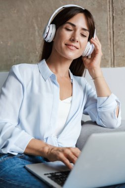 Evde rahat bir kadın, oturma odasında kanepeye oturup dizüstü bilgisayar kullanarak kulaklıkla müzik dinliyor..