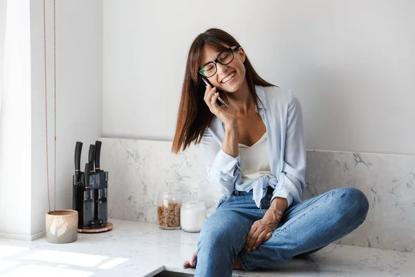 Joven feliz bonita mujer de negocios sentada en casa en la cocina hablando por teléfono móvil . — Foto de Stock