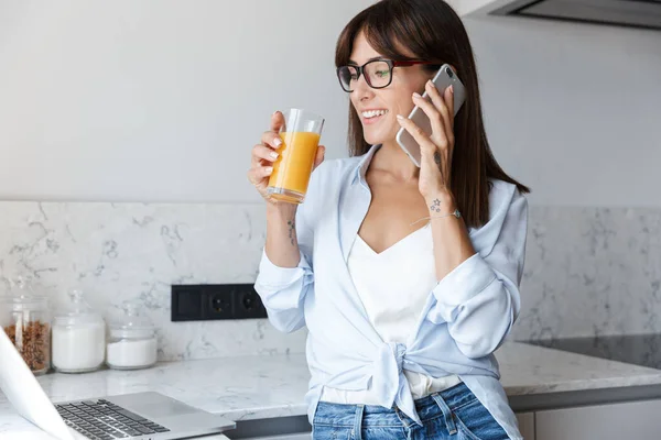 Задоволена бізнес-леді вдома на кухні, використовуючи ноутбук комп'ютер, що розмовляє мобільним телефоном, п'є сік . — стокове фото
