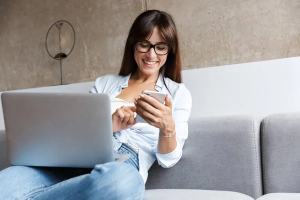 Gelukkig zakenvrouw binnen thuis op de bank in de woonkamer met behulp van laptop computer met behulp van mobiele telefoon. — Stockfoto