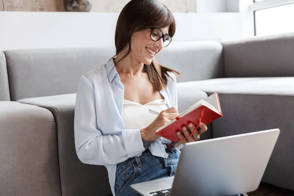 Positieve zakenvrouw binnen zitten op de vloer in de buurt van een bank in de woonkamer met behulp van laptop computer schrijven notities in notebook. — Stockfoto